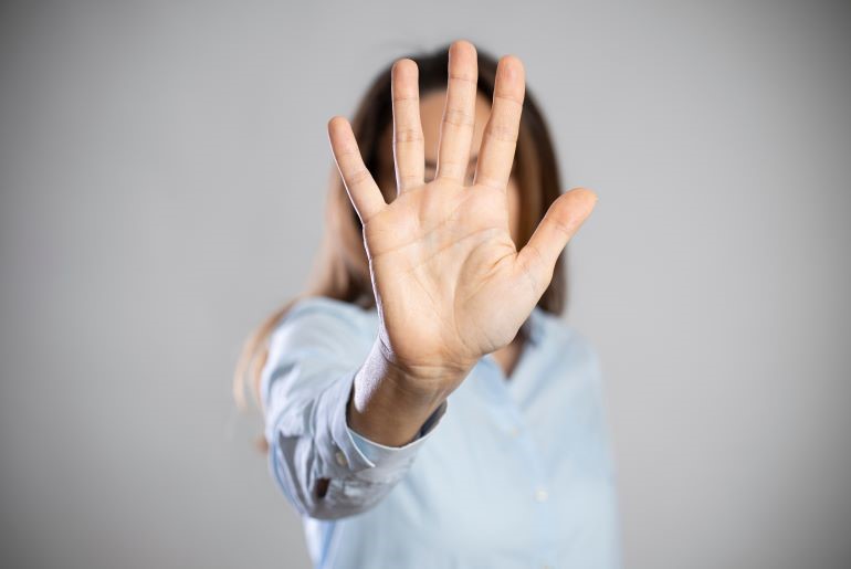 Abwehrend ausgestreckte Hand mit gespreizten Fingern vor hellblauem Oberteil, Gesicht verdeckt, grauer Hintergrund.