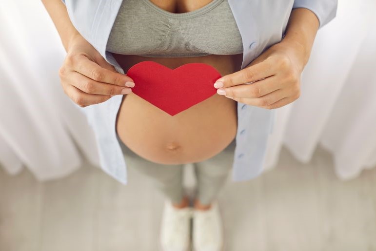 Schwangere Frau in weißem Hemd hält rotes Papierherz vor ihren Bauch.