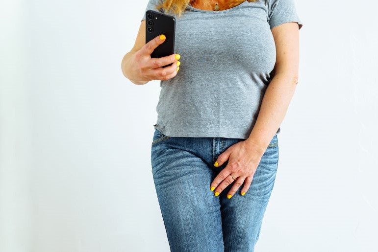 Frau in grauem T-Shirt und Jeans macht Selfie mit Smartphone. Beine aufgrund von Harndrang überkreuzt.