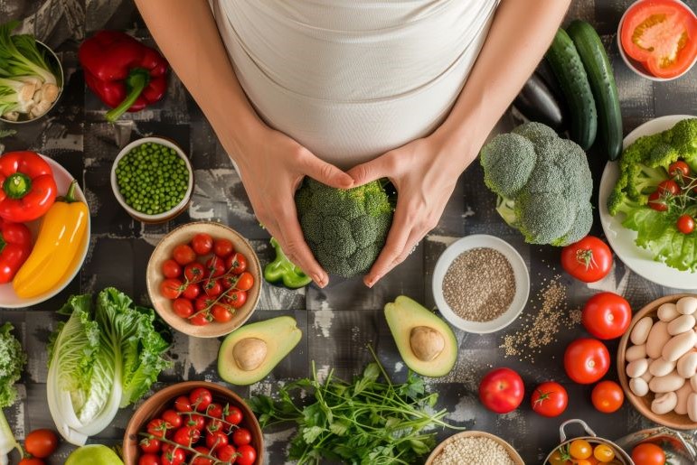 Schwangere formt Herz mit Brokkoli, umgeben von buntem Gemüse und Obst auf dunklem Tisch.