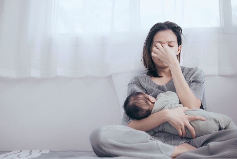 Erschöpfte Mutter mit Säugling auf Couch, Gesicht in Hand vergraben, zeigt peripartale psychische Belastung.