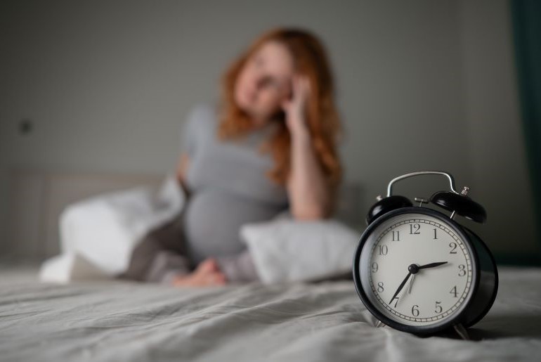 Schwangere Frau mit Schlafstörungen sitzt frustriert im Bett, Wecker steht im Vordergrund.