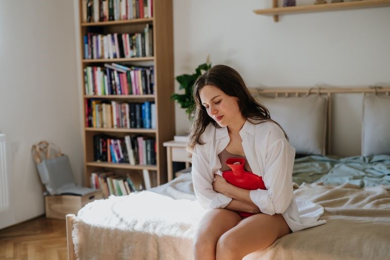 Frau mit Wärmflasche auf Bett zur Symptomlinderung bei Myomen und Endometriose. Bücherregal im Hintergrund.